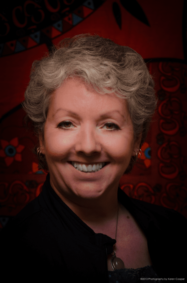 Smiling woman with short gray hair, wearing a black jacket against a bright yellow background, exuding warmth and approachability.