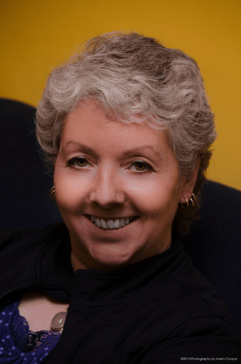 Smiling woman with short gray hair, wearing a black jacket against a bright yellow background, exuding warmth and approachability.