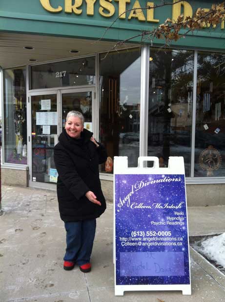 Smiling woman stands next to a sandwich board promoting "Angel Divinations" for psychic readings and Reiki, outside "Crystal Dreams" store.